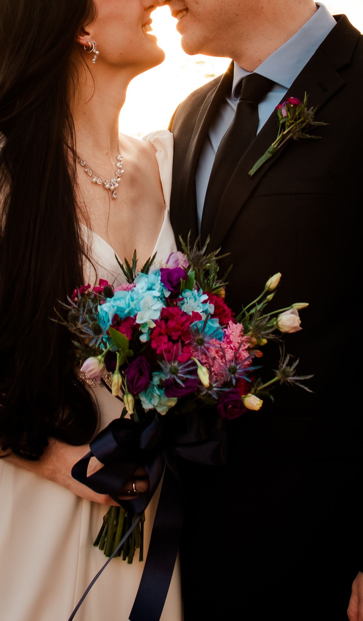 A close-up of a couple with a vibrant bouquet featuring blue and pink flowers at sunset.