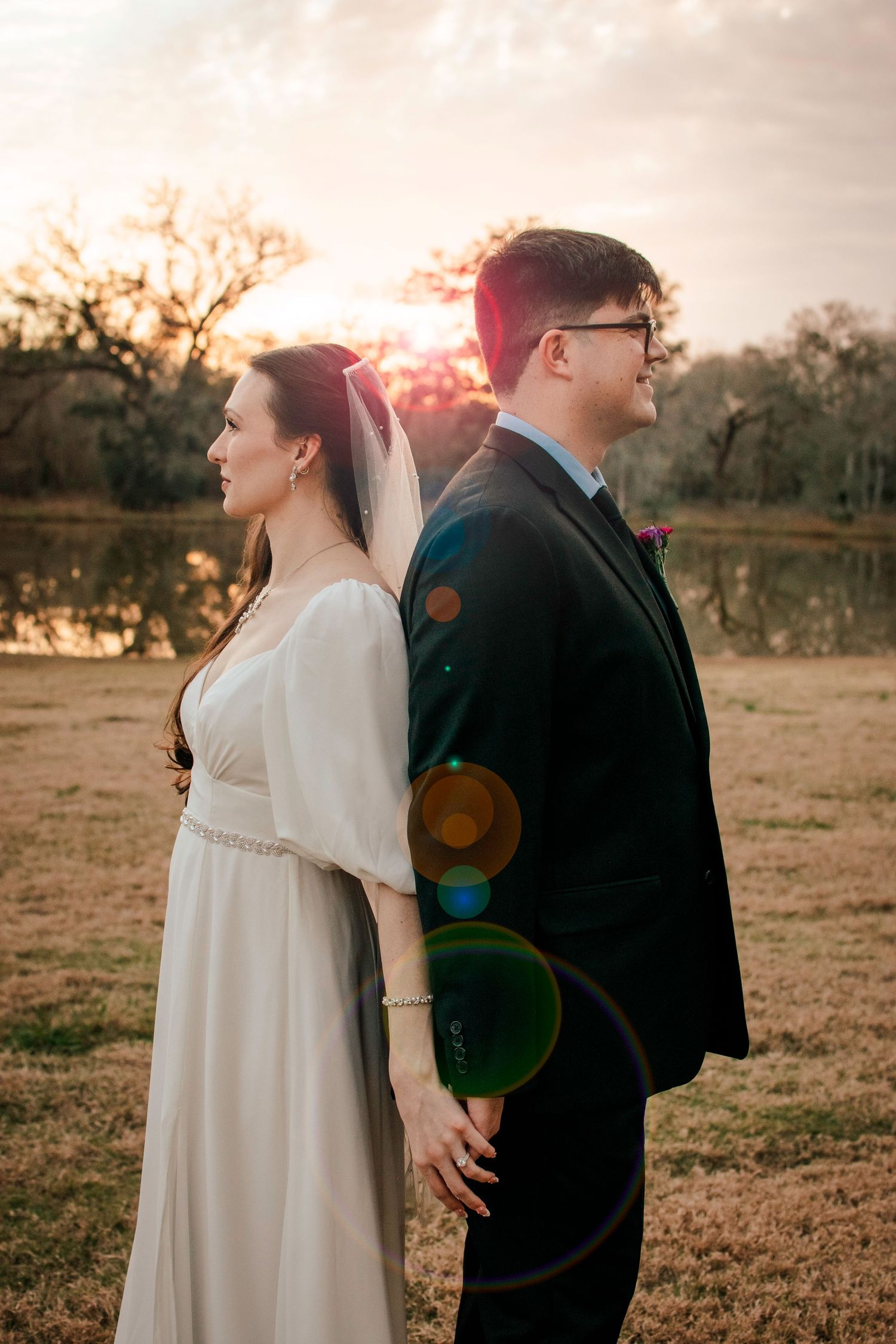 Romantic sunset wedding portrait with lens flare as newlyweds stand back-to-back by lake.
