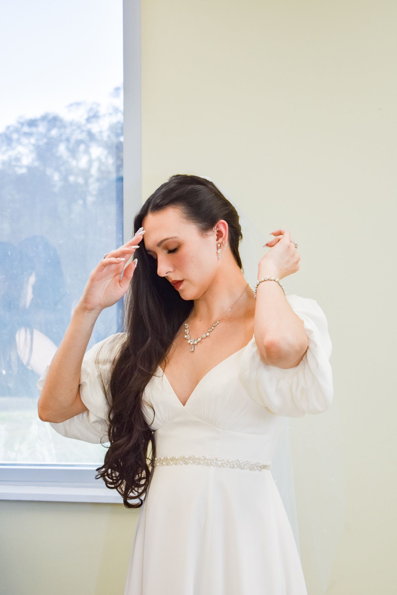 Elegant bride in white dress with long dark hair poses by window.