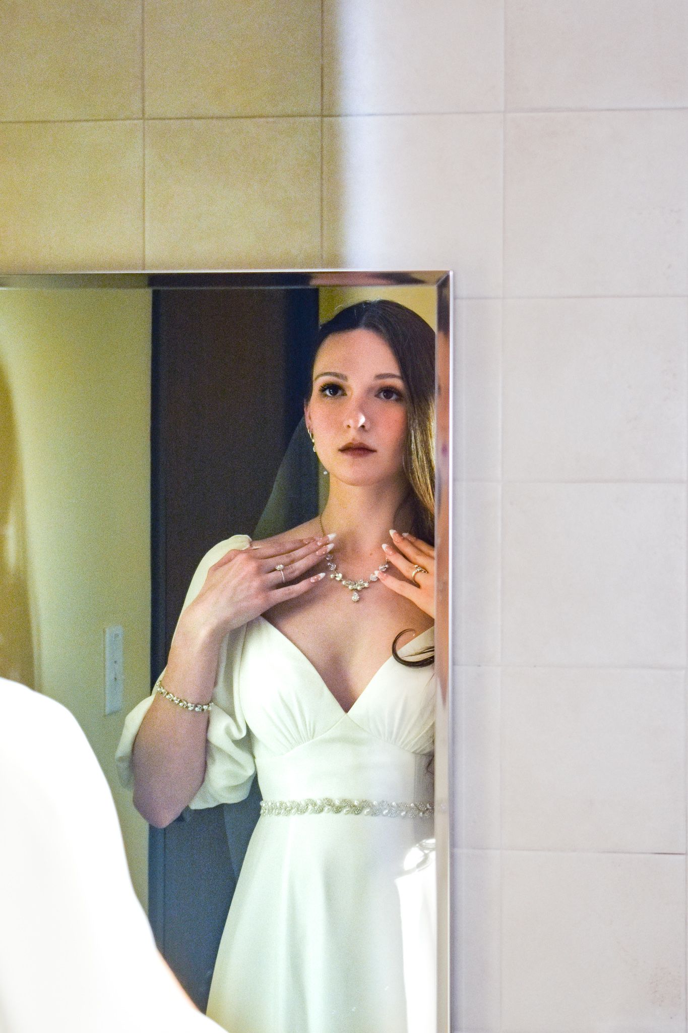 A bride in a white dress and jewelry looks at herself in a mirror while getting ready for her wedding.