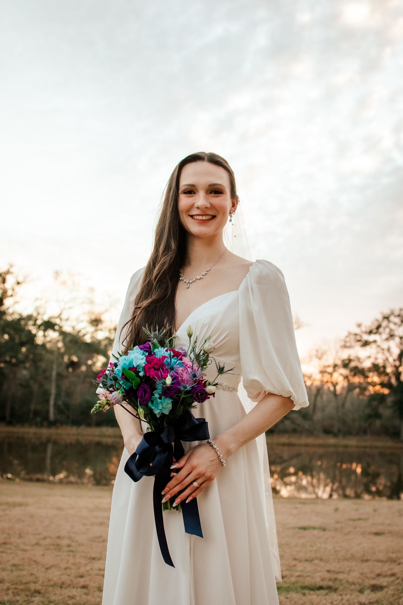 Bride in flowing white dress holds vibrant blue and purple bouquet during sunset portrait session.