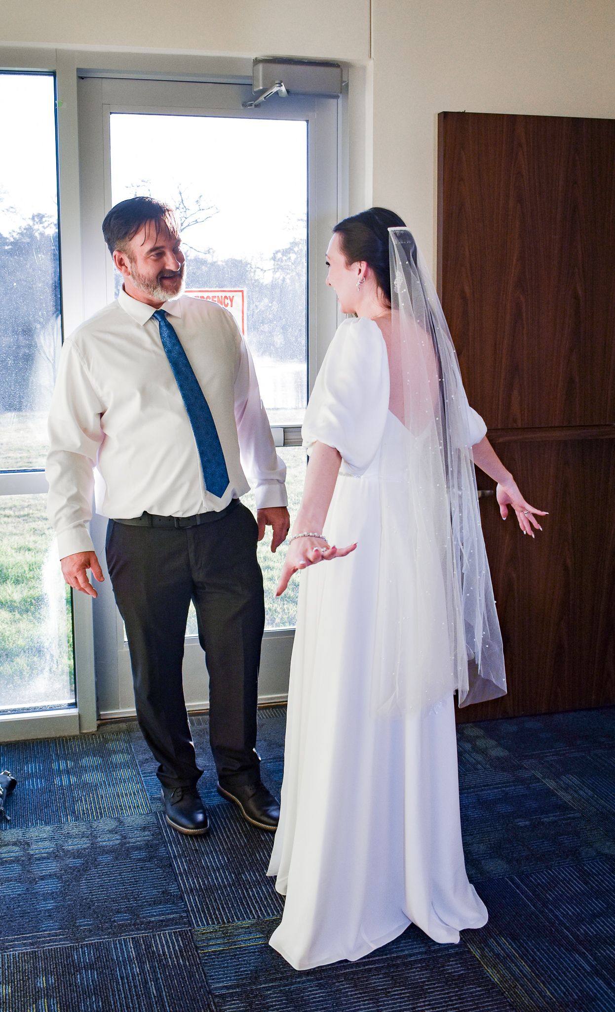 A couple in wedding attire shares a candid moment in front of a window.