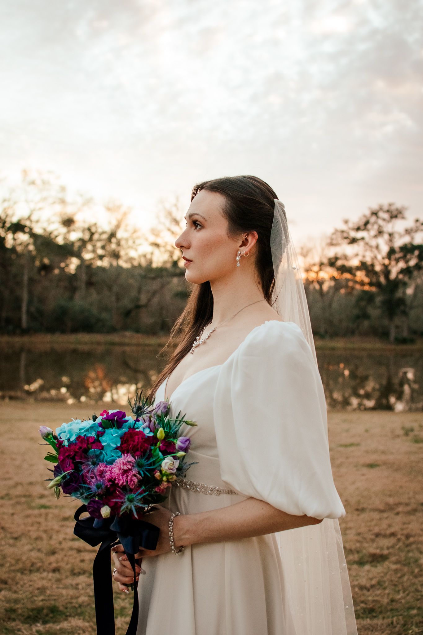 Side profile of bride holding colorful bouquet against sunset sky and lake backdrop.