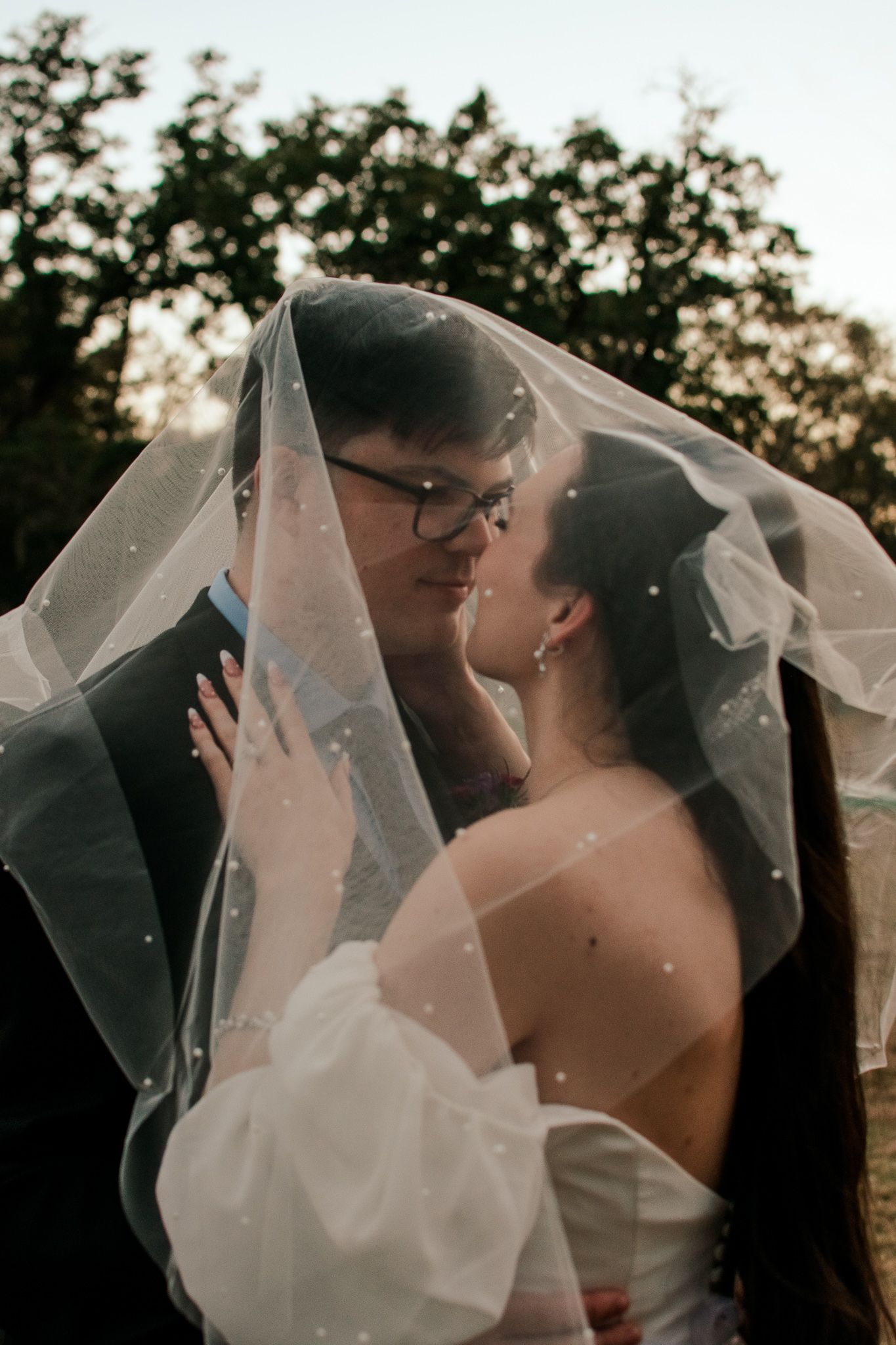 Artistic wedding portrait of couple beneath flowing veil at sunset.