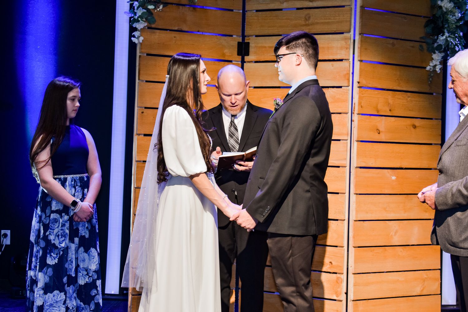 A wedding ceremony takes place on stage with wooden decor and blue lighting.