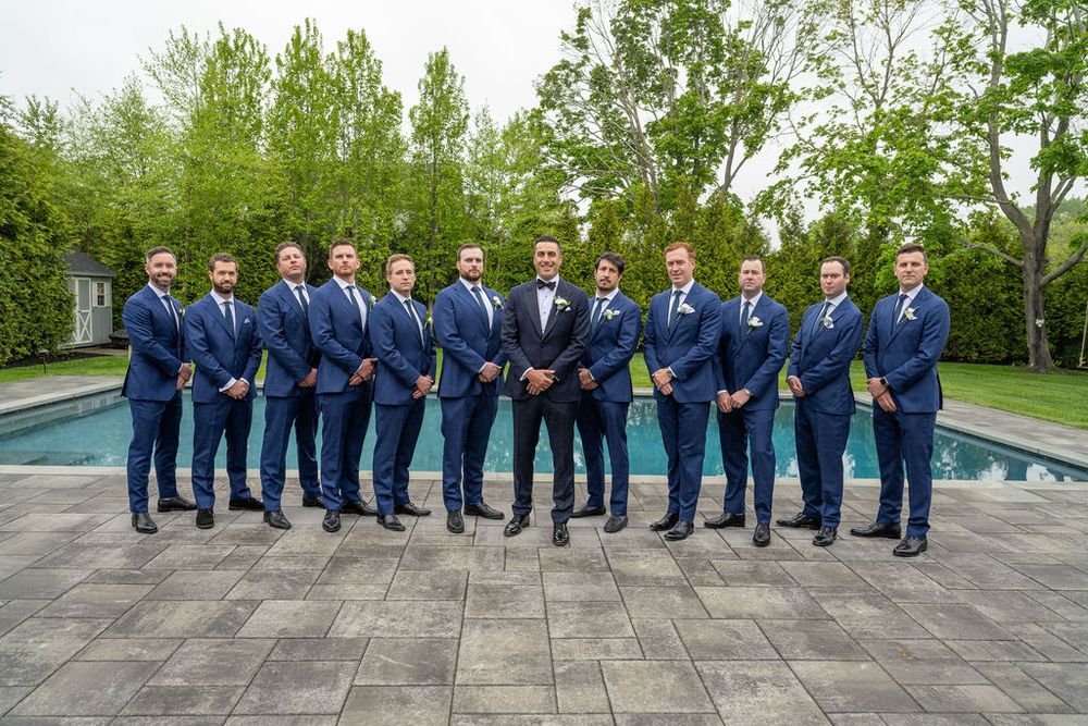 Groomsmen in matching navy suits posing formally by poolside surrounded by lush green trees.