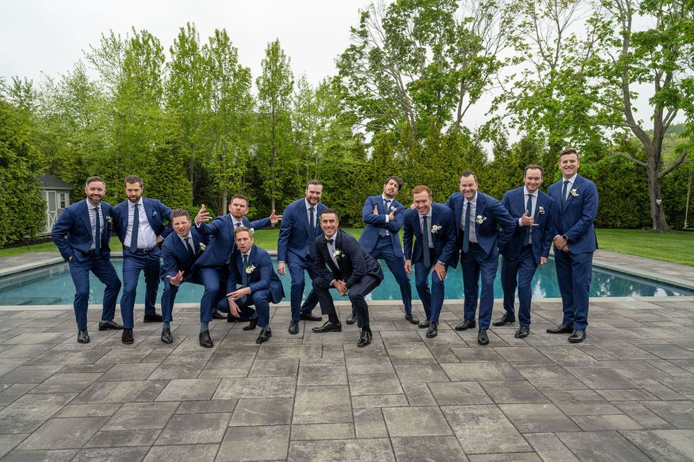 Fun groomsmen group photo in navy suits striking playful poses by outdoor pool with green trees.