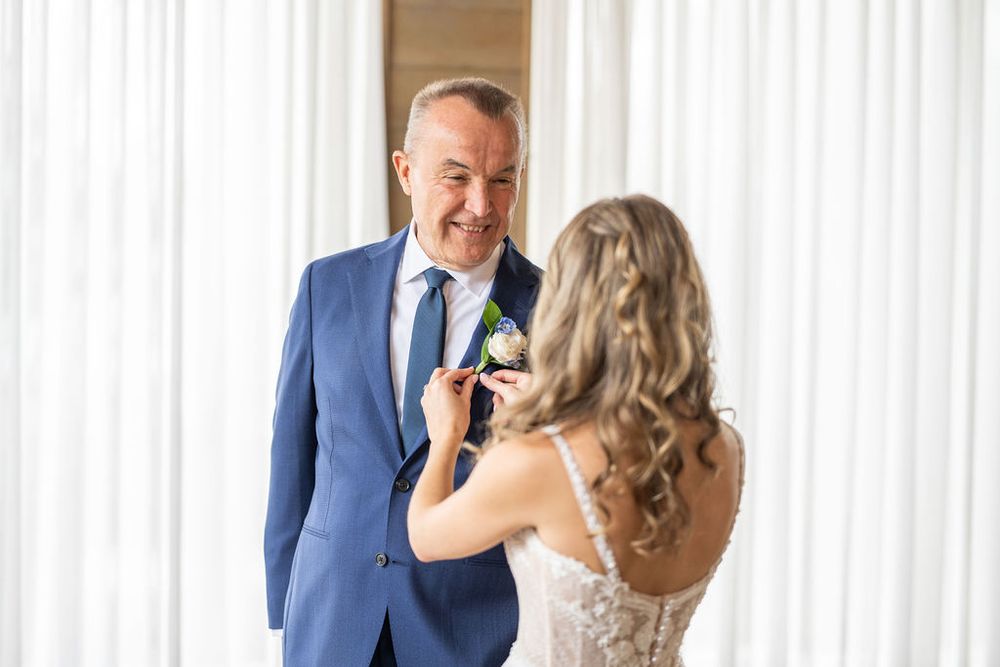 Bride pinning boutonniere on smiling groom in navy suit during wedding preparation indoors.