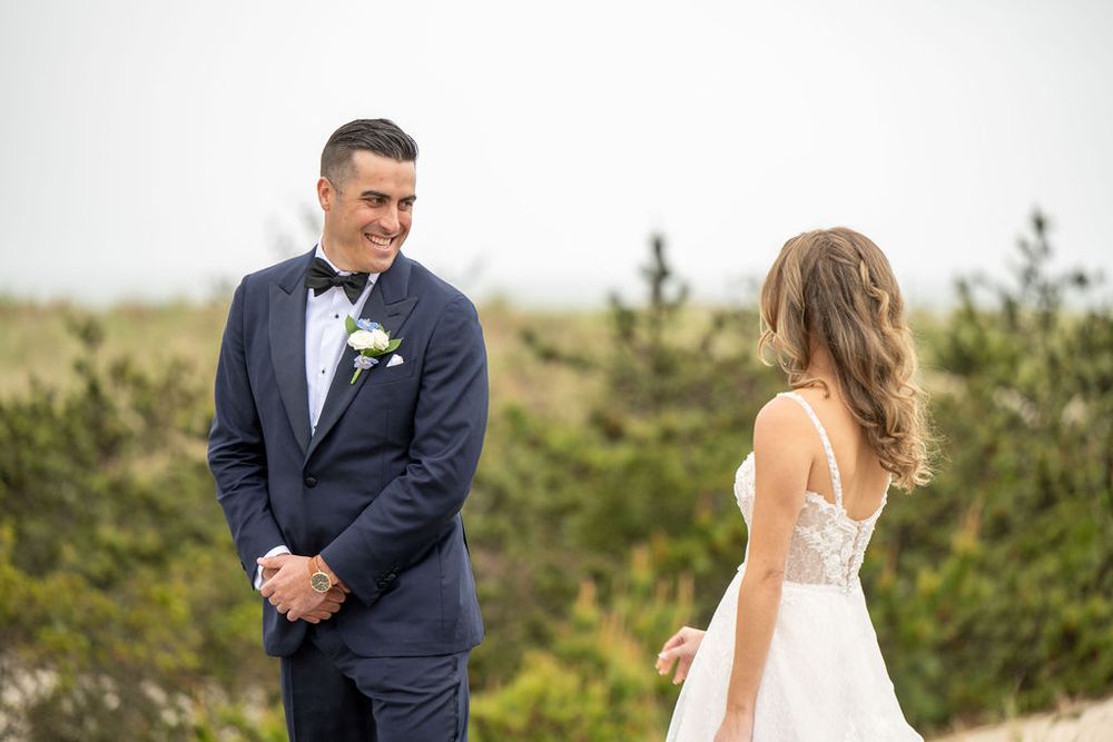 Emotional first look moment as groom in navy tuxedo smiles at bride among natural dune landscape.