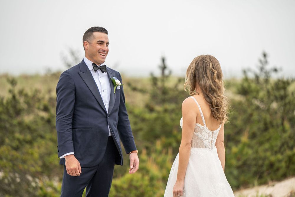 Groom in navy tuxedo smiles at bride in lace wedding dress during outdoor first look near sandy dunes.