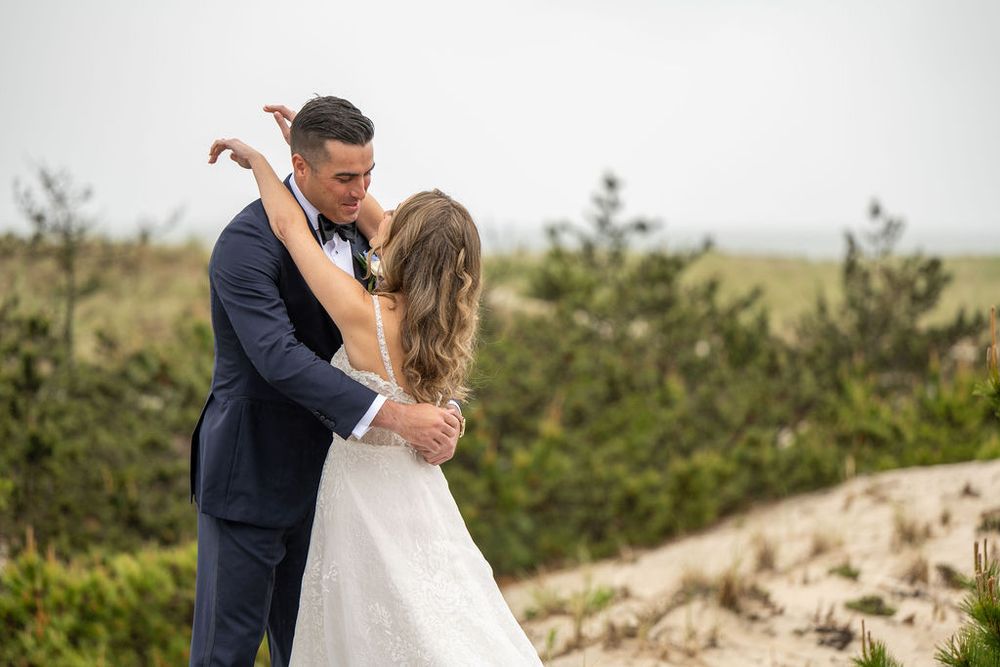 Groom in navy suit embraces bride in elegant lace gown on sandy beach dunes during wedding photos.
