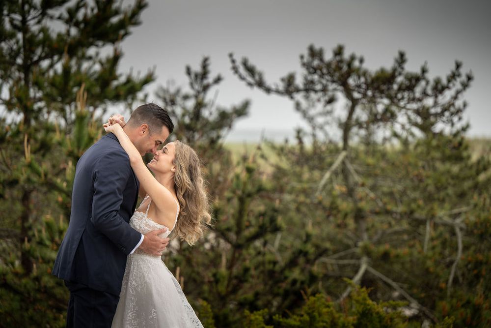Bride and groom share a romantic kiss surrounded by lush green pine trees on their wedding day.