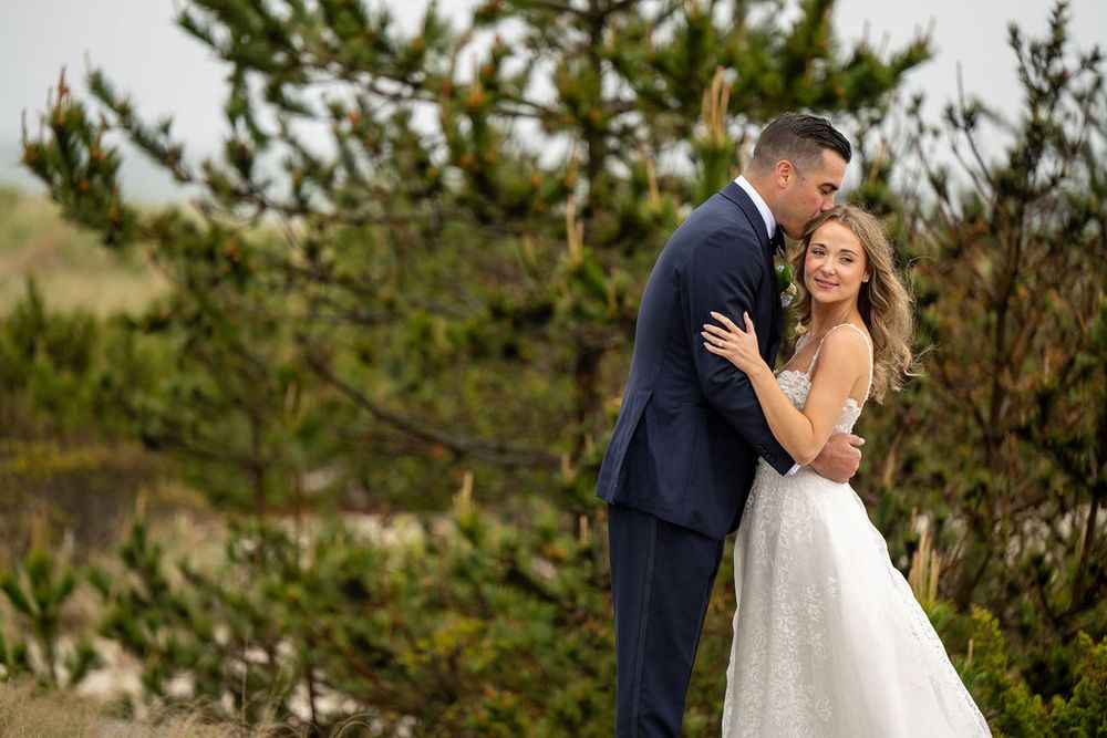 Smiling bride in lace gown leans into groom in navy suit while posing near pine trees on wedding day.