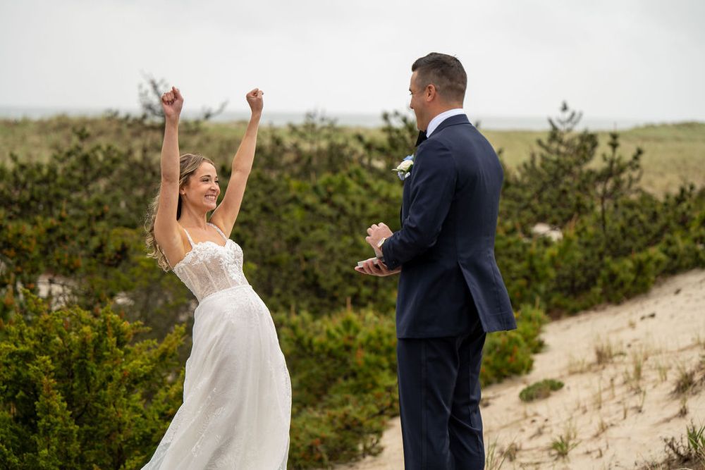 Excited bride raises arms in joy during first look reveal with groom on sandy beach wedding location.