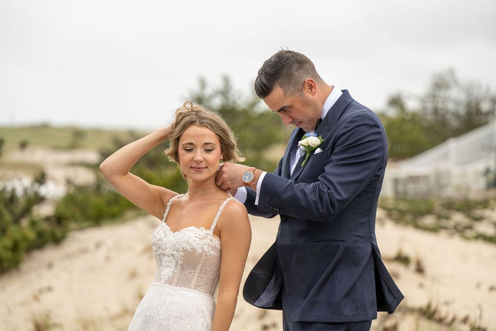 Groom in navy tuxedo fastens a delicate necklace on bride in lace gown during outdoor wedding photos.