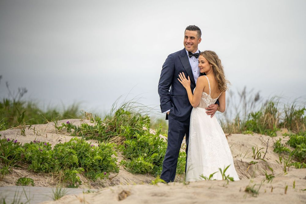 Bride and groom pose romantically on sandy beach dunes with lush green coastal grass in the background.
