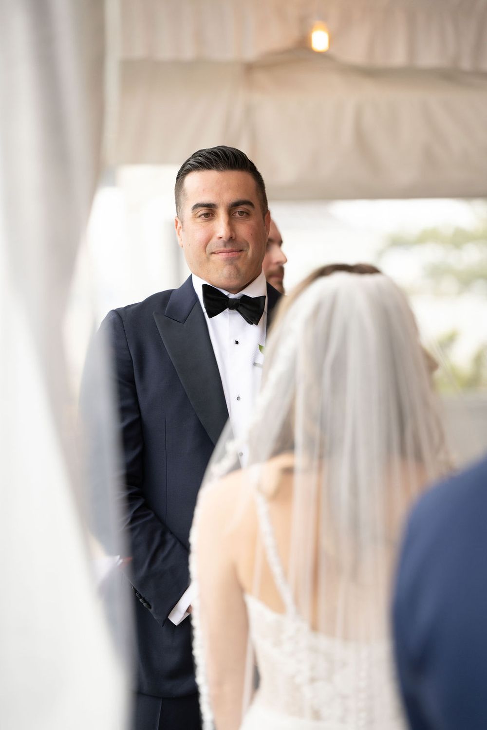 Emotional groom in black bow tie watches bride approach during outdoor tented wedding ceremony.