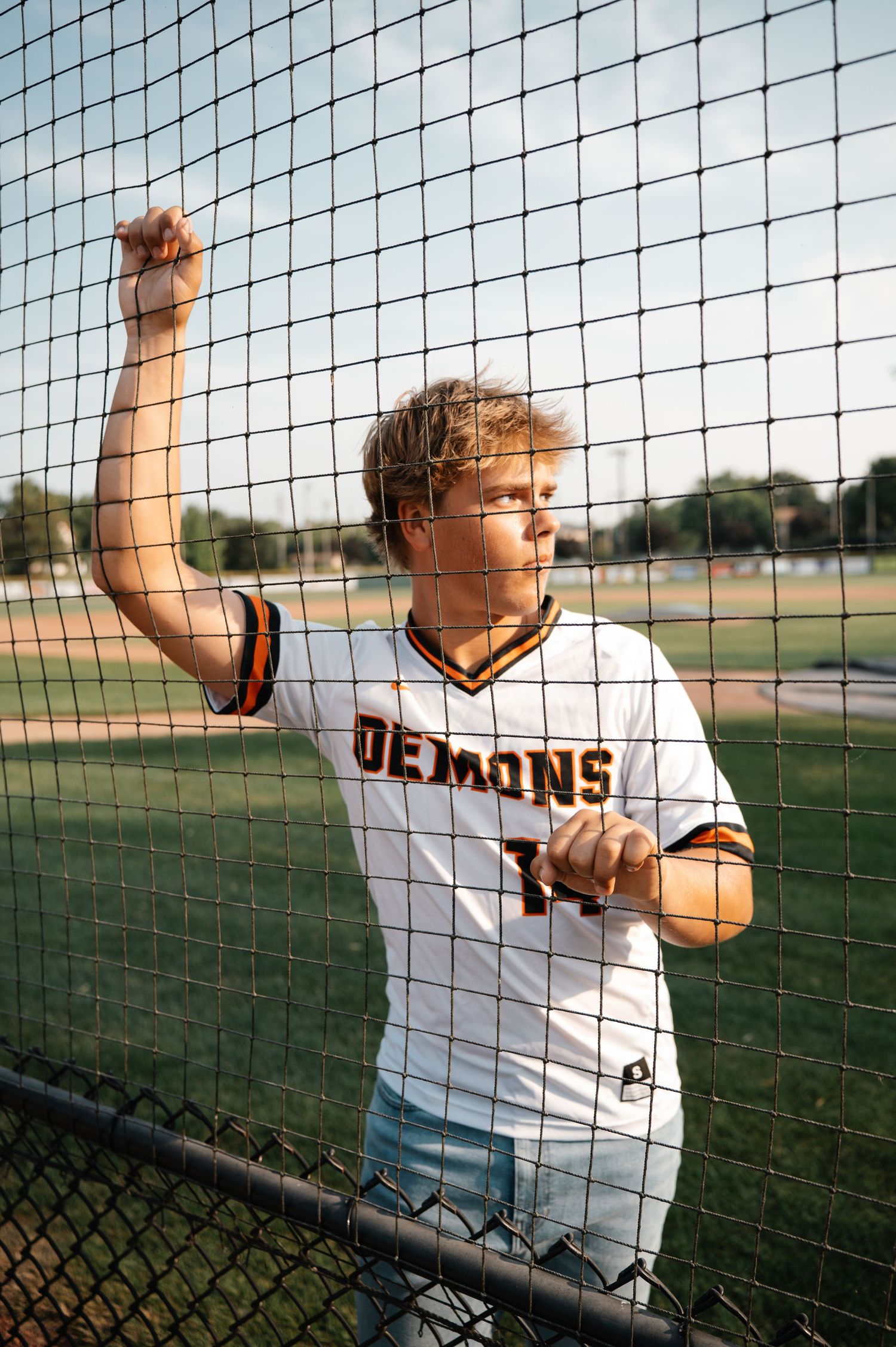 Teen baseball player in Demons jersey leaning against chain-link fence at outdoor field.
