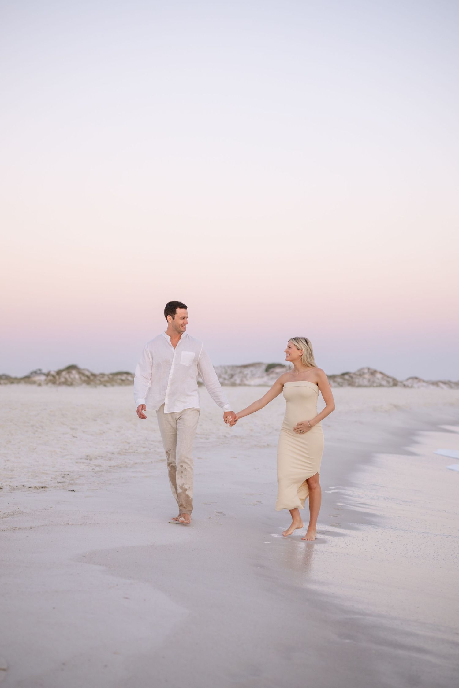 A couple walks together along the shoreline at sunset in neutral colored outfits.