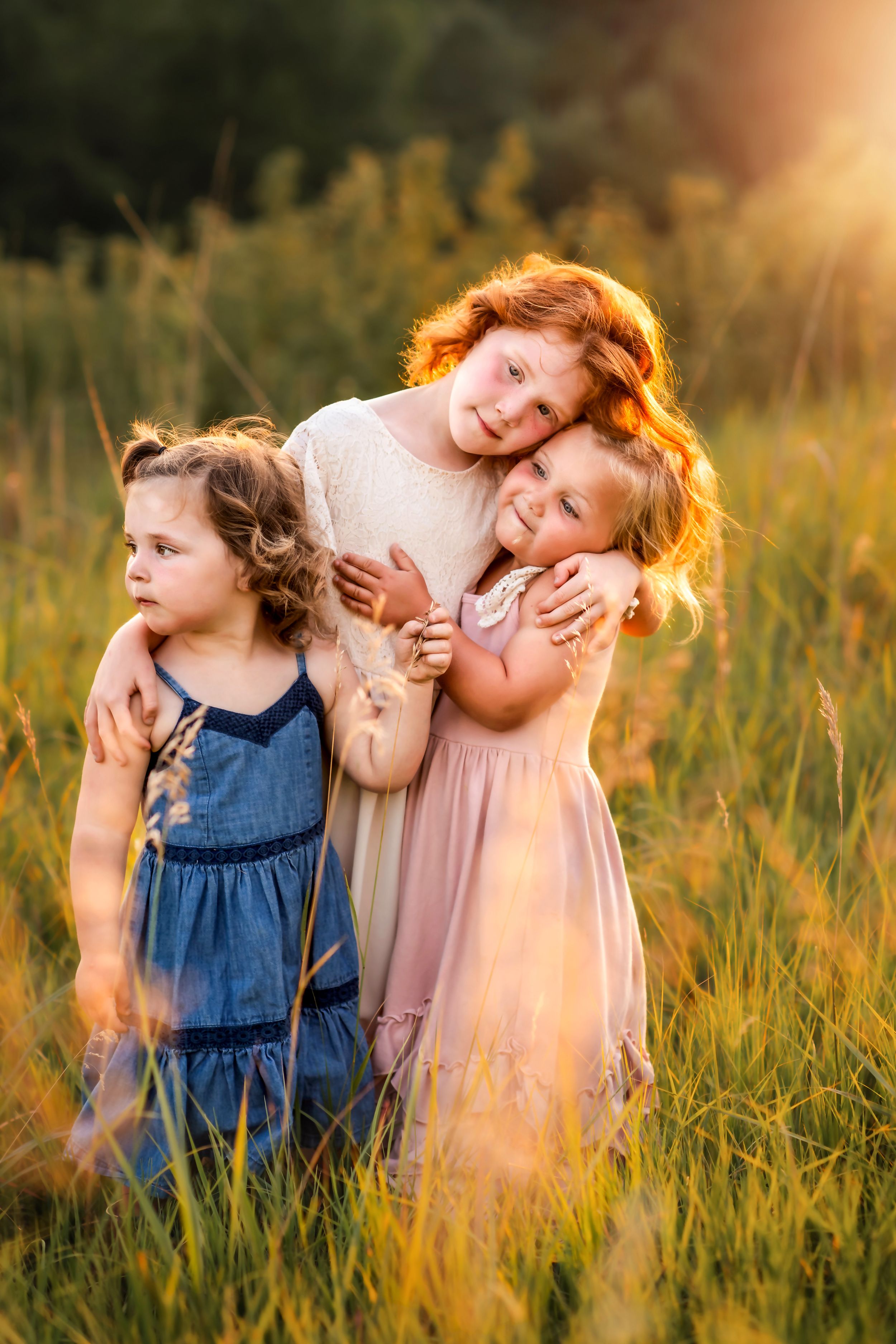 Three young children hugging and laughing together in a sunlit meadow at sunset.
