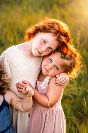 Two children in light colored dresses embrace while smiling in golden sunlit field.