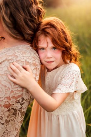 Child with red curly hair wearing white lace dress hugs someone in matching outfit outdoors.