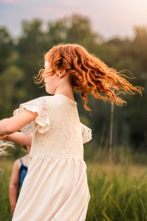Child in white dress twirls with flowing red hair in sunlit meadow at dusk.