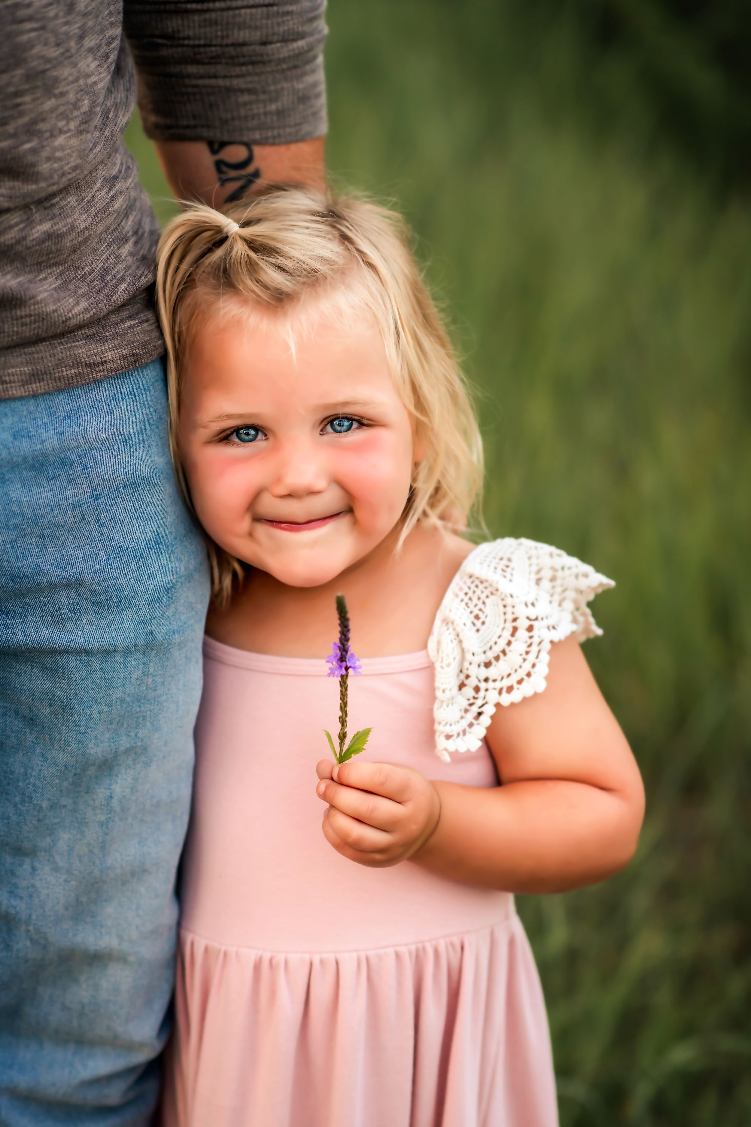 Small child in pink dress holds purple flower while standing next to someone in casual wear.