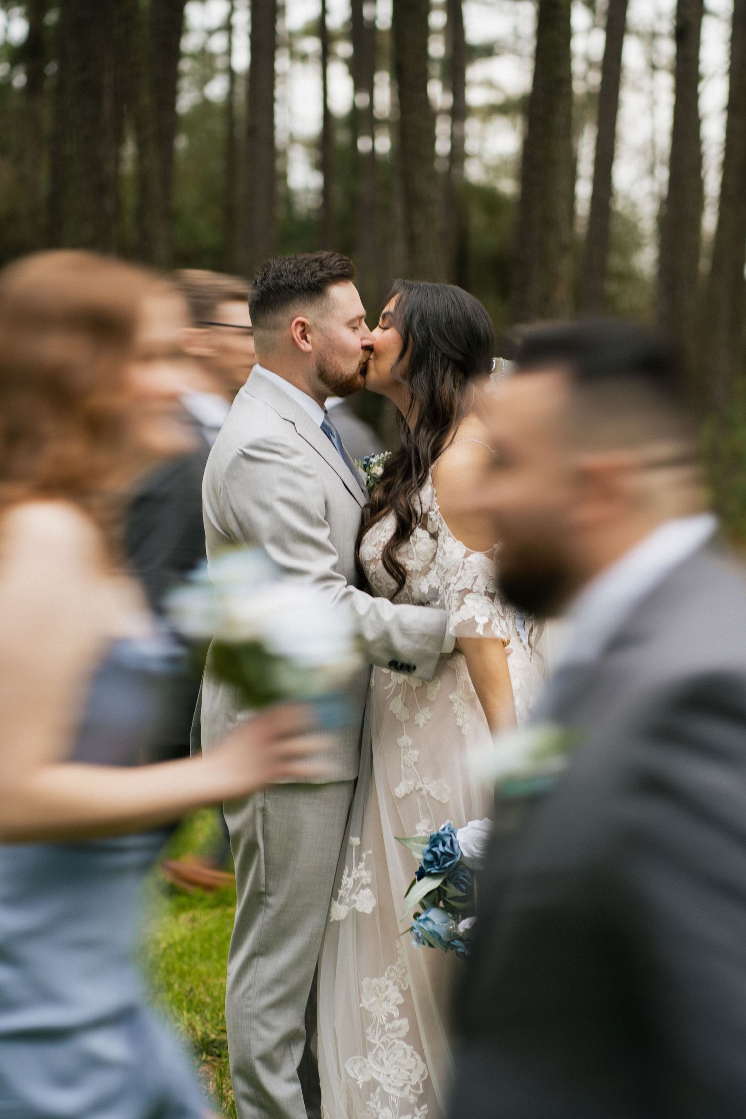 A wedding ceremony moment captured with the couple kissing while guests look on in a wooded setting.