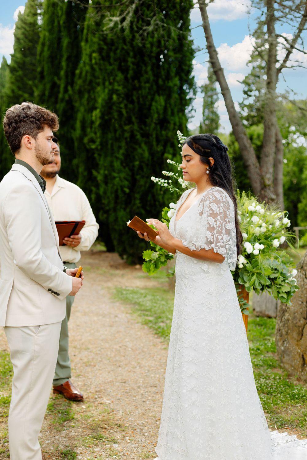 Outdoor wedding celebration in a sunlit garden pathway lined with cypress trees.
