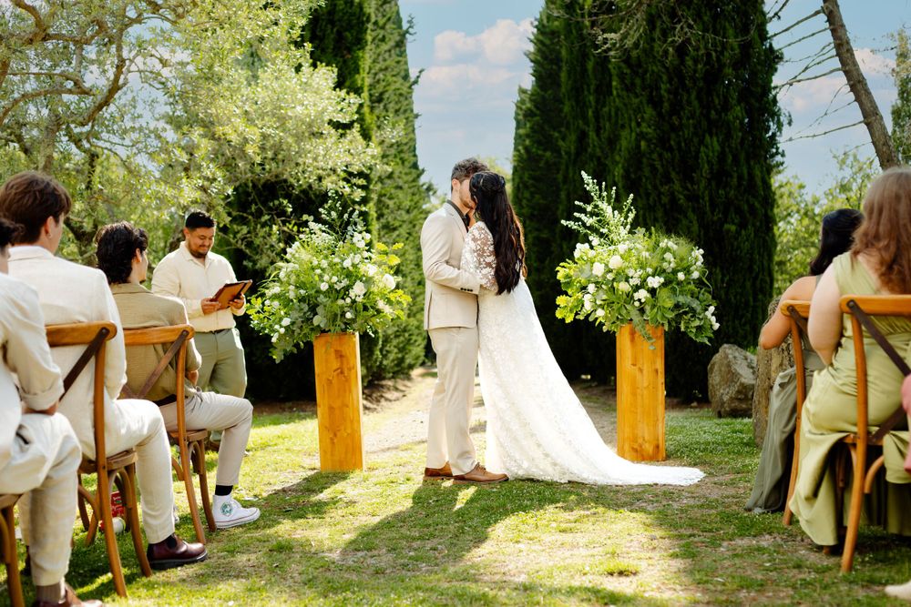 Wedding guests seated in wooden chairs for an outdoor garden ceremony under tall trees.
