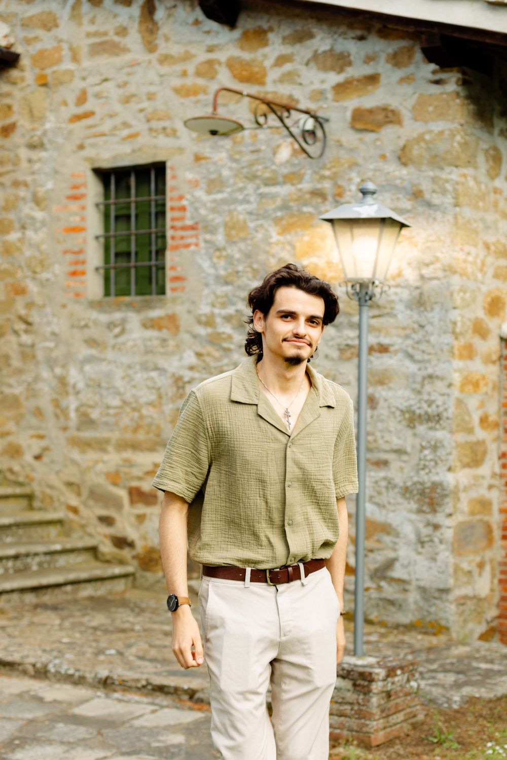 Person in vintage-style attire walking near stone wall with window and lantern.