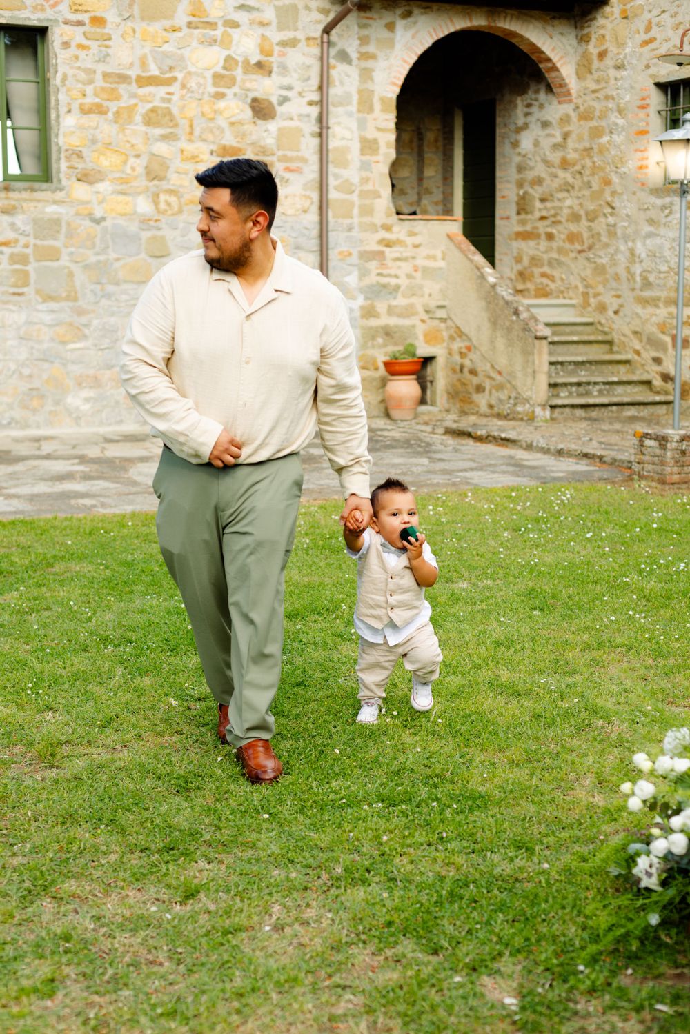 Father and toddler walking together on grass in front of rustic stone building.