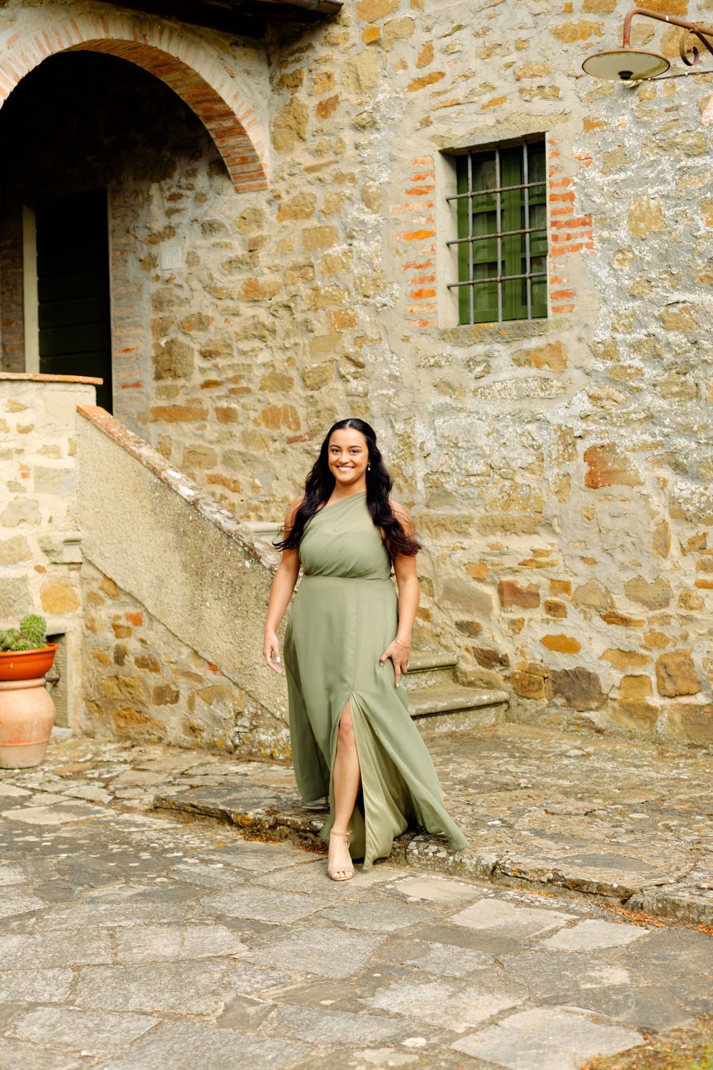 Woman in elegant olive green dress posing by stone wall with arched doorway.