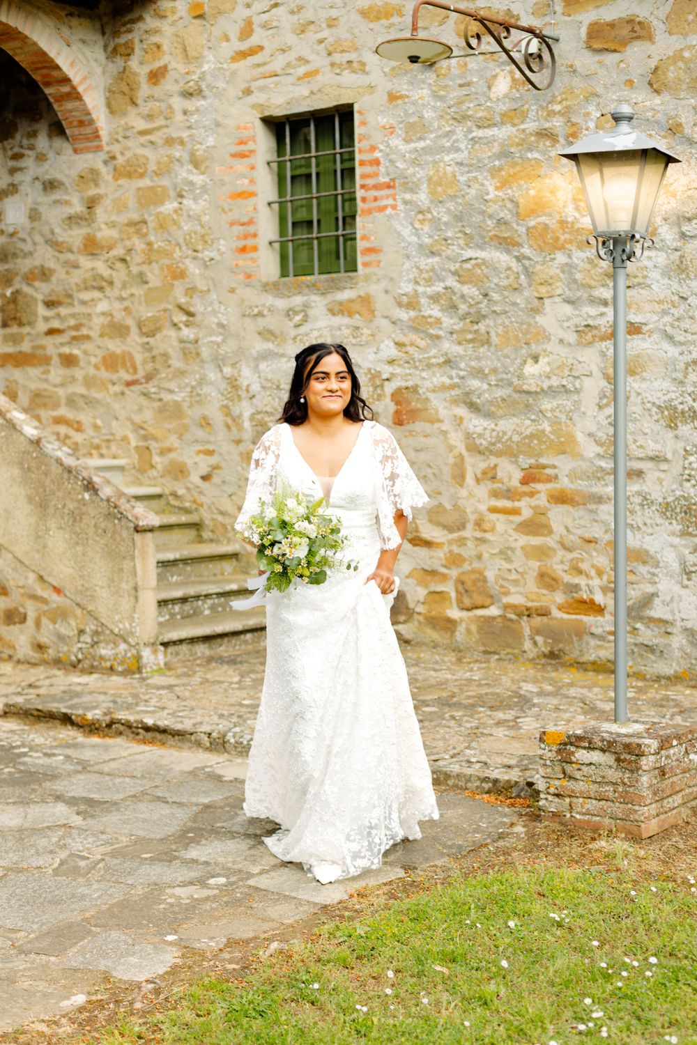 Bride in white lace dress holding bouquet stands by rustic stone building with stairs.