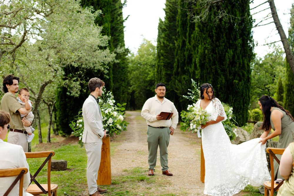 Outdoor wedding ceremony with guests seated on benches under cypress trees.