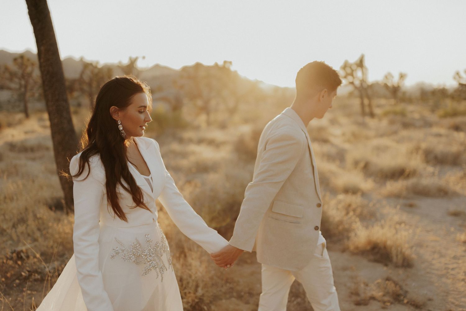 A couple in white walking hand in hand through a desert landscape at golden hour.