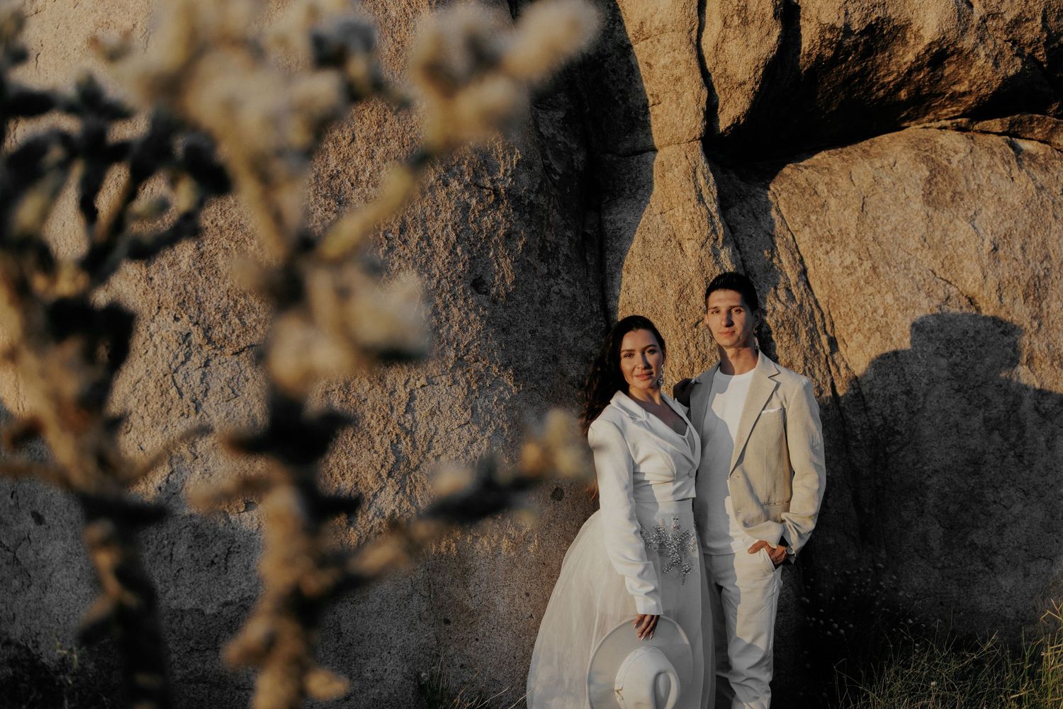 A couple in white attire poses against a rocky desert backdrop during golden hour.