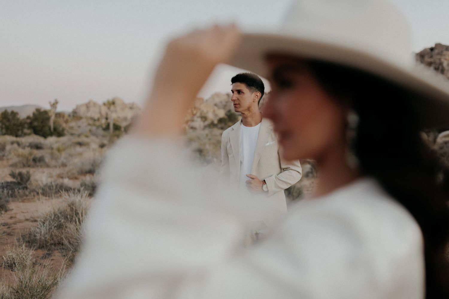 A stylish couple in all-white outfits poses together in a desert landscape during golden hour.