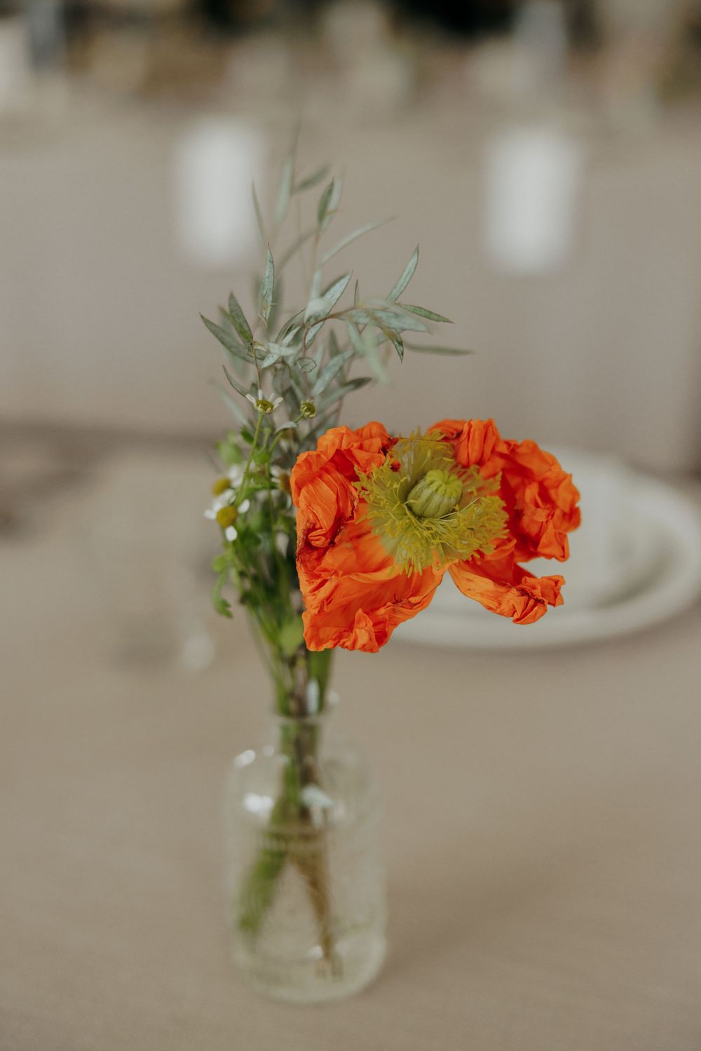 Orange poppy flower arrangement with olive branches in clear glass vase.