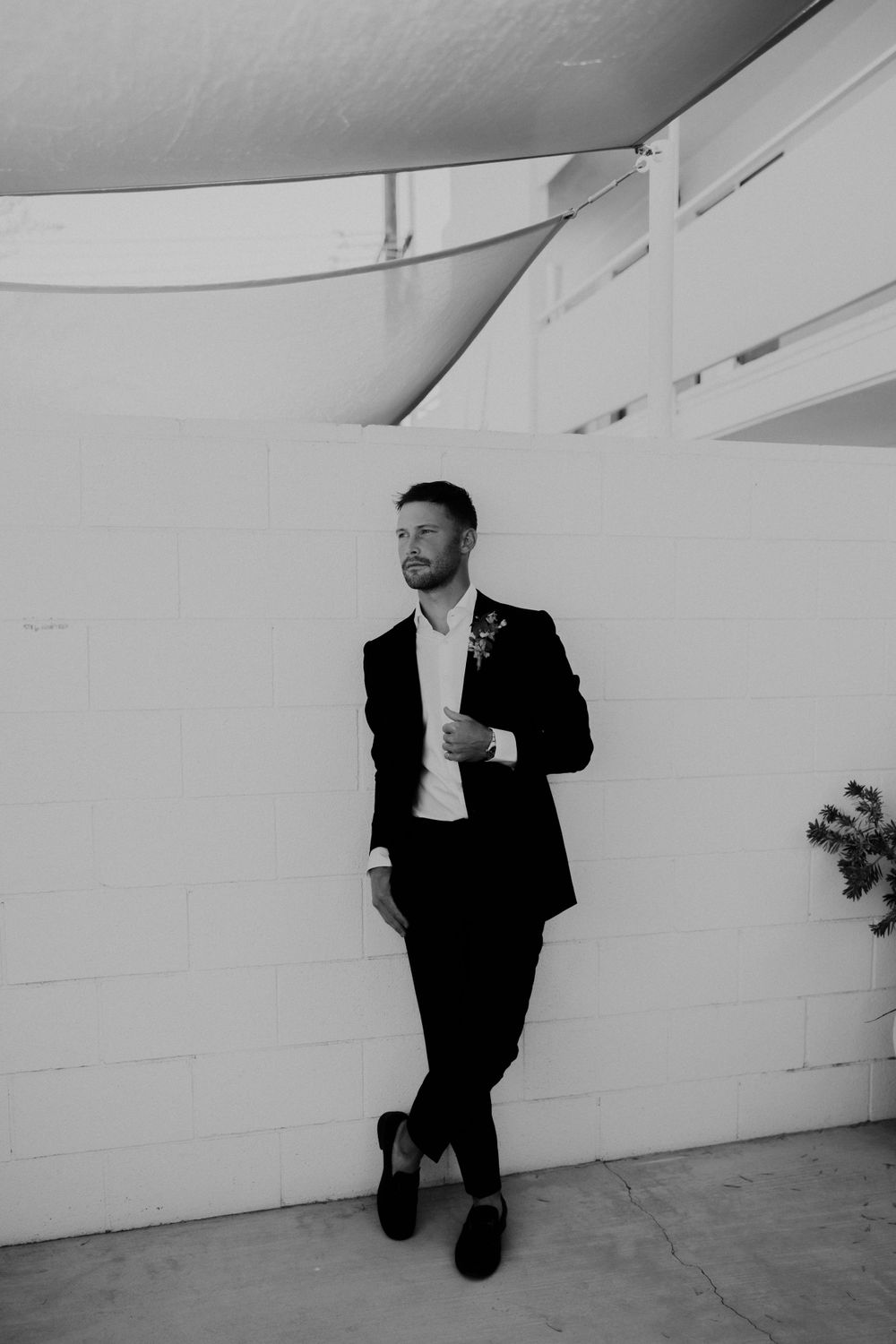 Black and white portrait of groom leaning against white wall in formal attire.