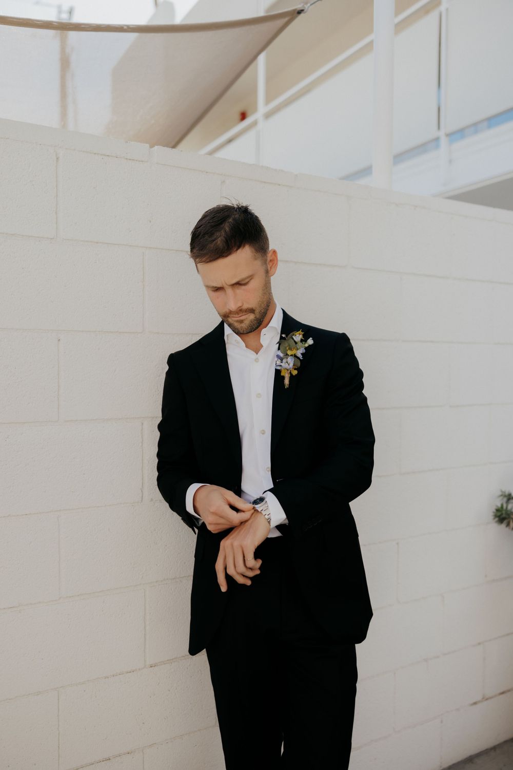 A groom in a black suit and white shirt checks his watch while standing against a white wall.