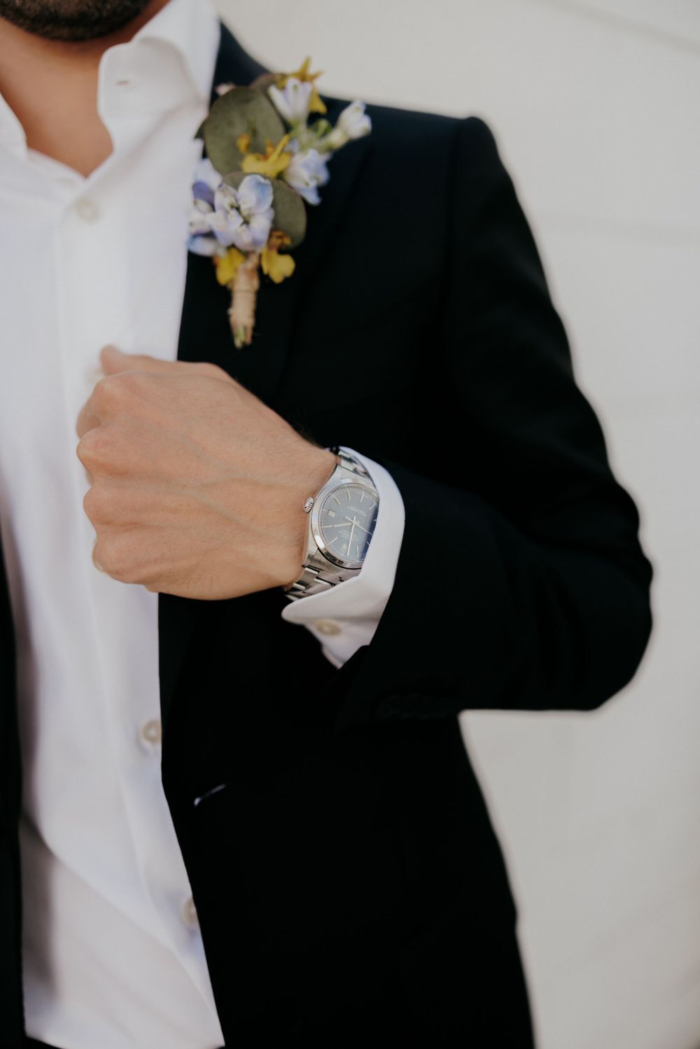 Close-up of a silver watch and boutonniere on a formal black suit jacket.