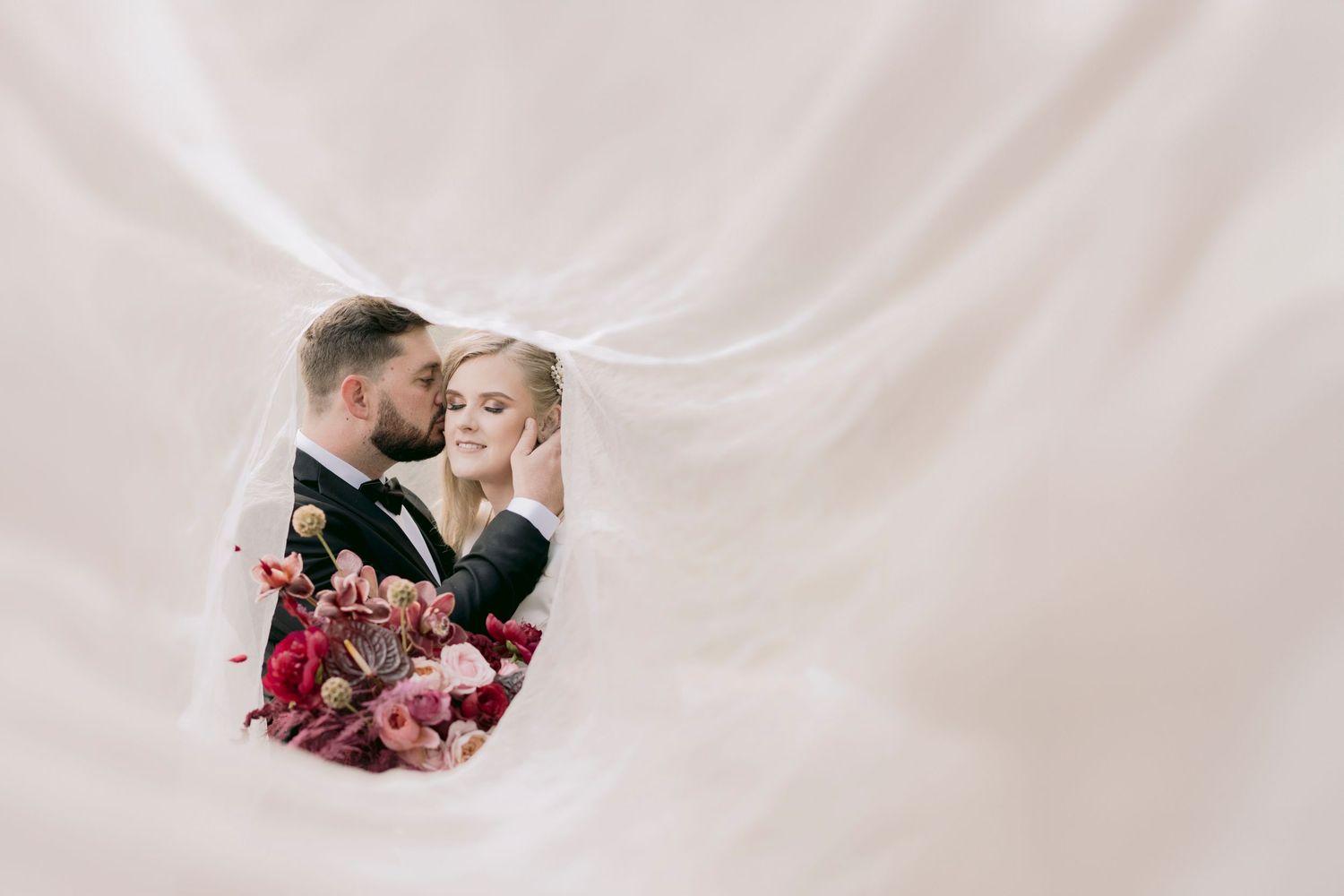 Romantic wedding couple embracing tenderly framed by a flowing white bridal veil.