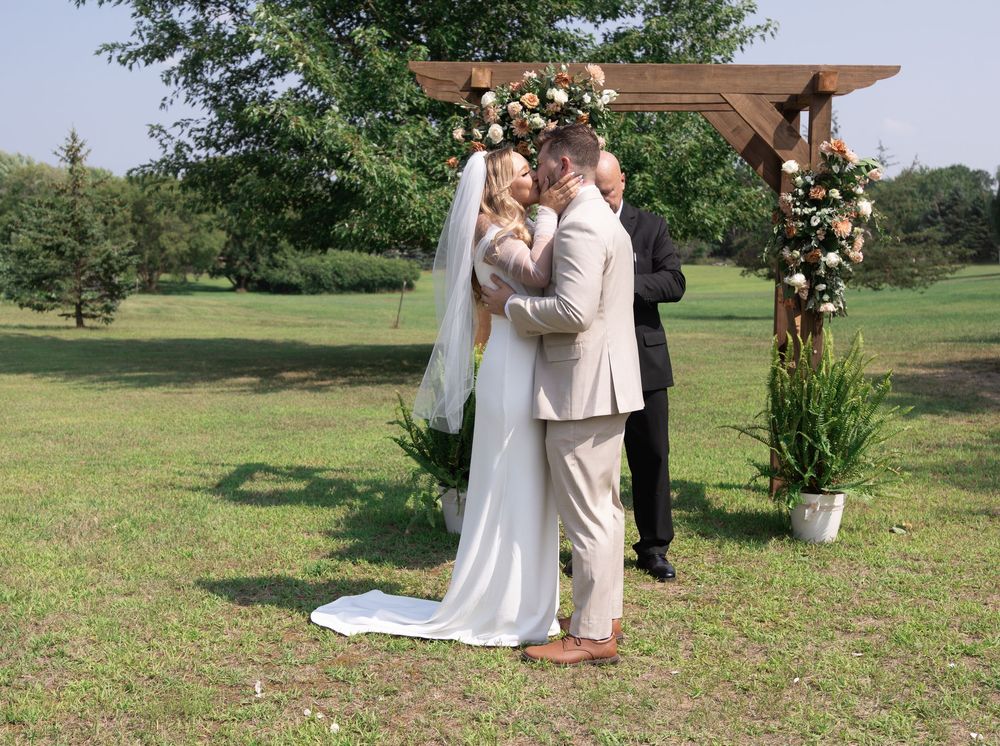 Romantic outdoor wedding ceremony at a rustic venue with a wooden arbor adorned with peach flowers on a grassy field.