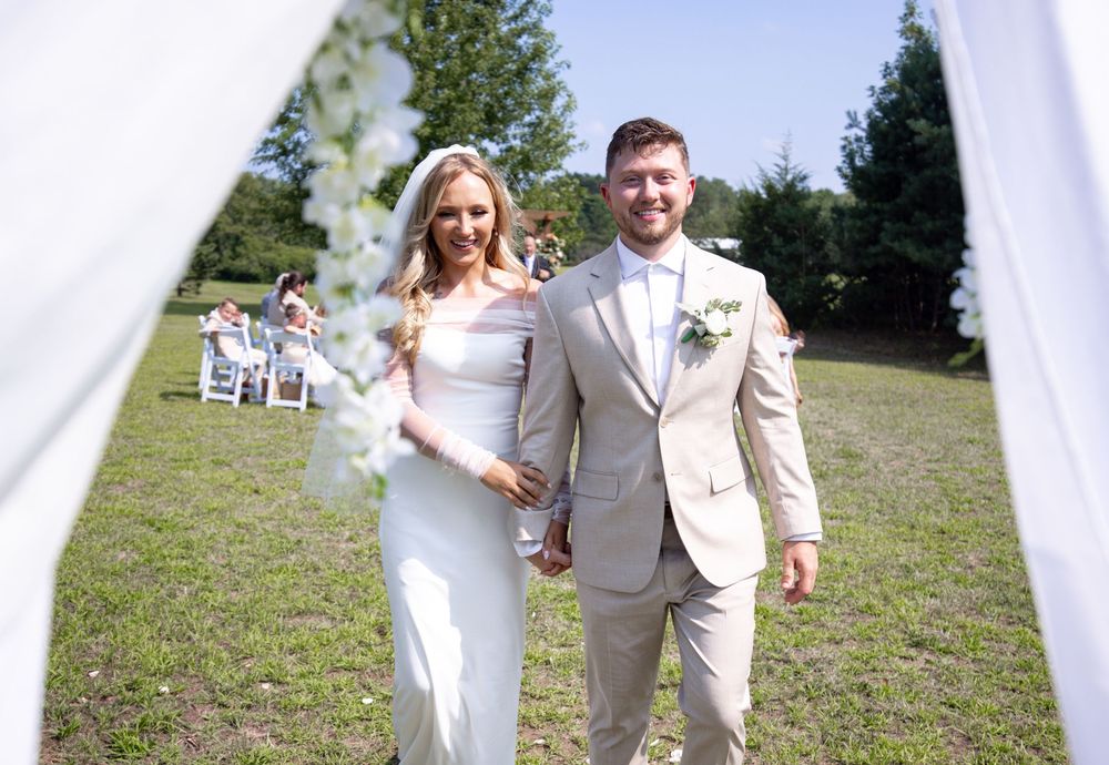 Happy couple walking with their wedding party on a grassy field with white draping decor.