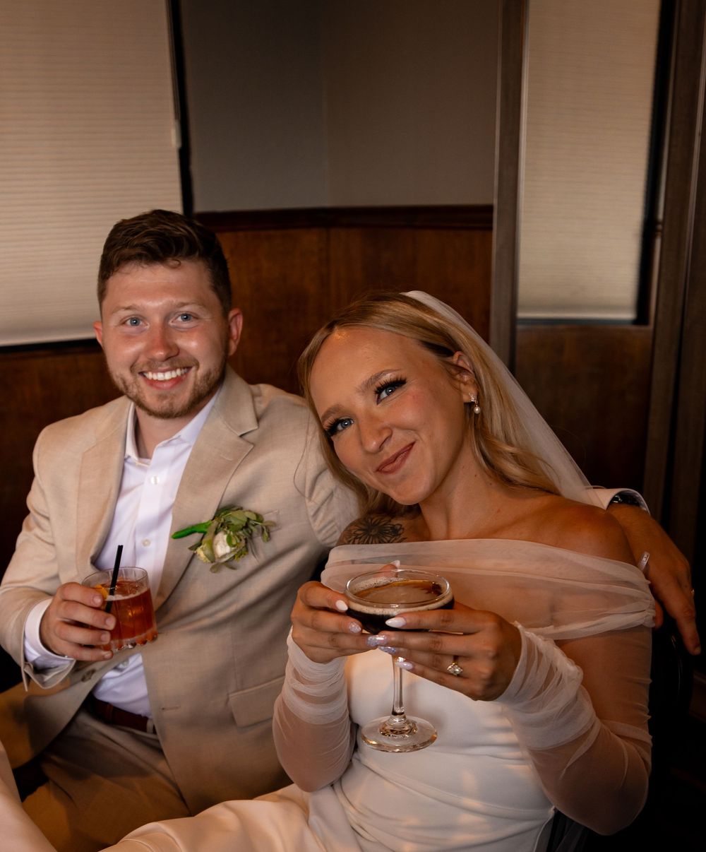 Newlywed couple smiles while holding wine glasses during wedding reception at indoor venue.