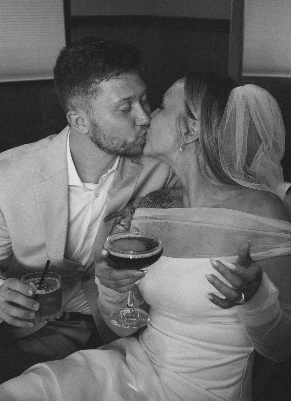 Black and white photo of bride and groom sharing romantic kiss while holding drinks.