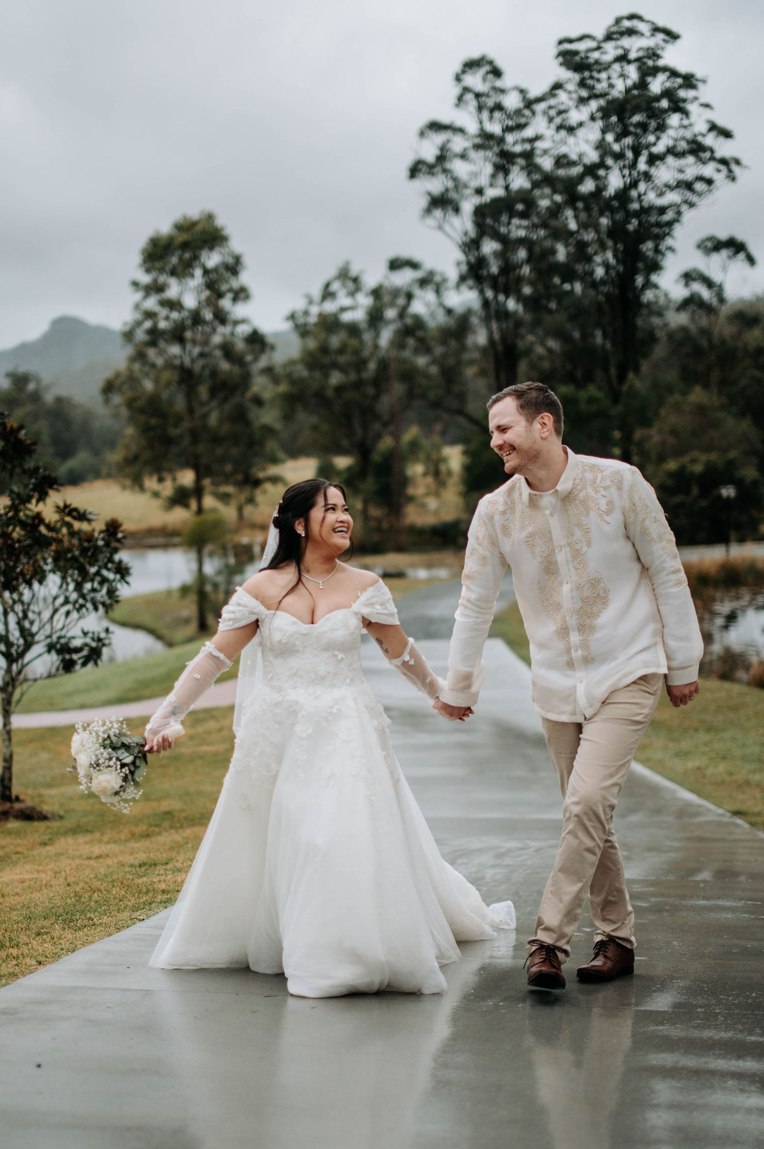 Joyful wedding couple walking on wet path with mountain landscape backdrop.
