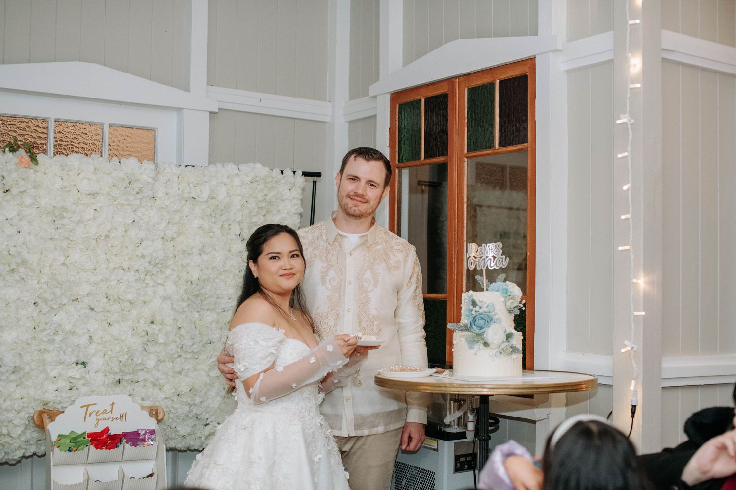 Newlyweds stand by their wedding cake against white floral backdrop during reception.